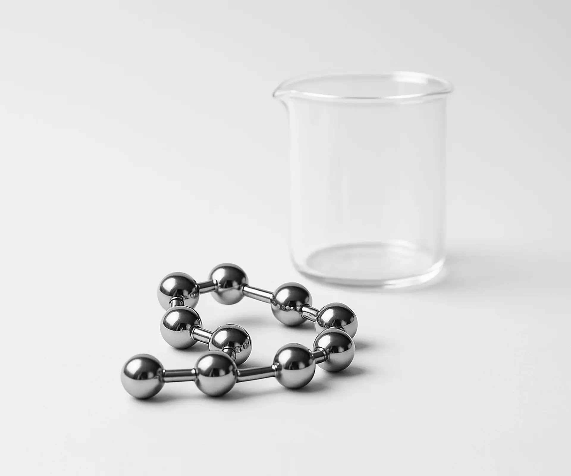 Chrome ball-and-stick molecular chain model beside a clear glass beaker on a white bench