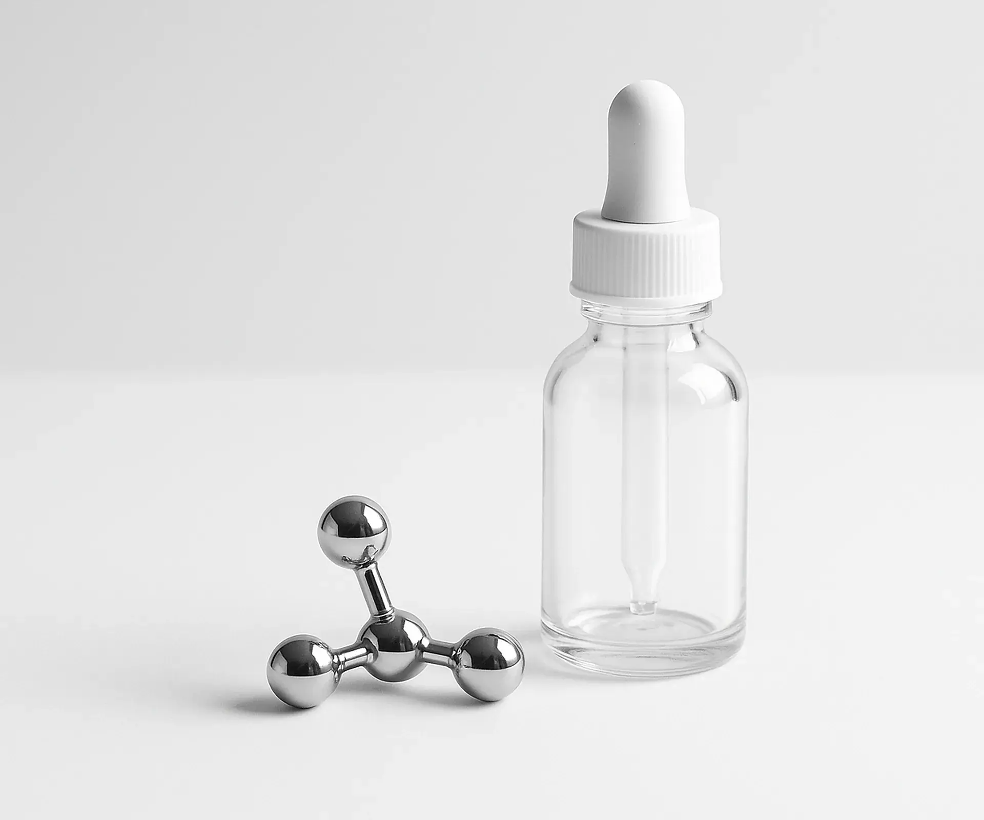Small chrome ball-and-stick molecule model beside a clear glass dropper bottle on a white bench